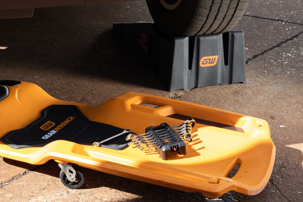 Automotive workspace shot showing an orange Gearwrench rolling mechanic creeper and a black under-car plastic wheel ramp supporting a vehicle tire, with a modular hand wrench set organized on the creeper surface.