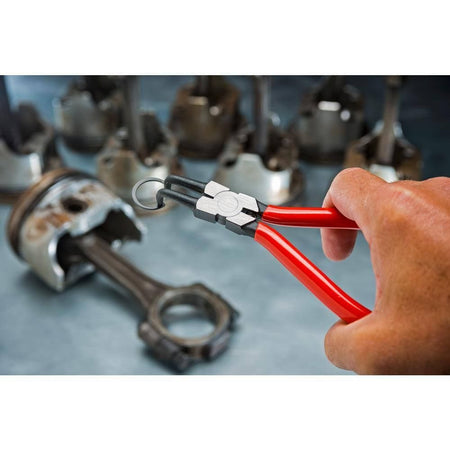 Action shot of a technician holding the red-handled Gearwrench internal snap ring pliers to compress and install a metal retaining ring inside an automotive engine piston assembly on a workshop table.
