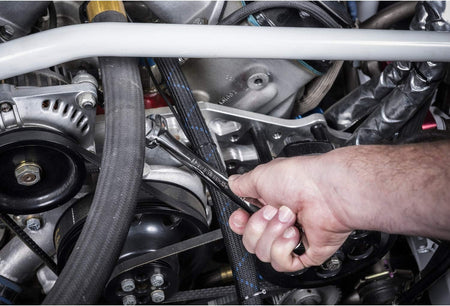 Action perspective of a field technician utilizing the open-end configuration of a long-pattern Gearwrench alloy steel spanner to tighten an adjustable fluid component fitting underneath an engine frame link.