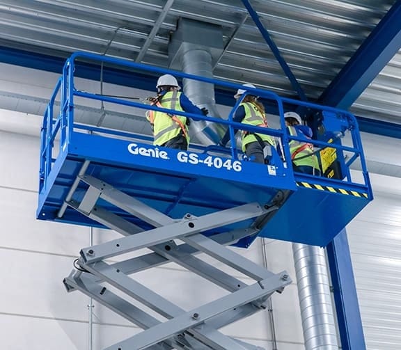 Three workers on extended Genie GS-4046 scissor lift inspecting ductwork in an industrial building with metal ceiling panels.