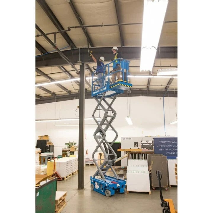 Two workers wearing safety helmets and harnesses stand on a fully extended Genie GS-1930 scissor lift platform inside an industrial facility. They are inspecting or working on ceiling infrastructure, demonstrating safe elevated access for maintenance tasks in a manufacturing or warehouse environment.