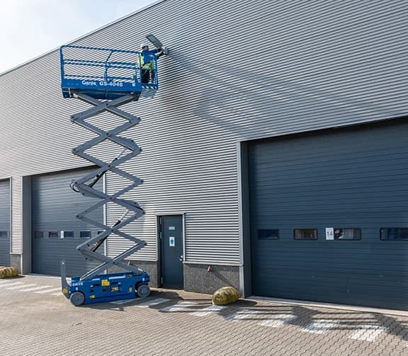 Genie GS-4046 scissor lift fully extended with a worker accessing the upper wall of a large industrial building. The lift is positioned on a paved surface near roll-up doors and yellow safety barriers, demonstrating elevated access for exterior maintenance tasks.