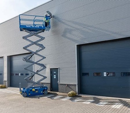 Genie GS-4046 scissor lift fully extended with a worker accessing the upper wall of a large industrial building. The lift is positioned on a paved surface near roll-up doors and yellow safety barriers, demonstrating elevated access for exterior maintenance tasks.