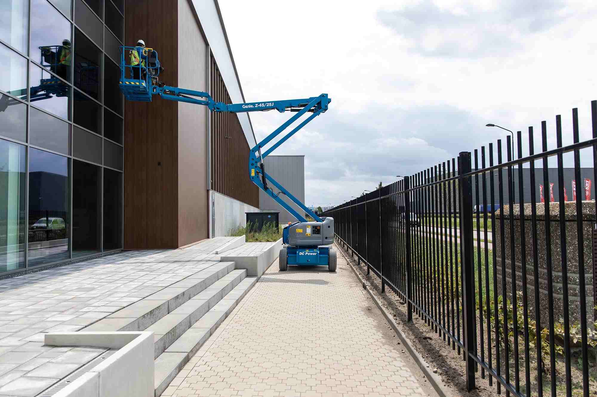 Genie Z-45/25J boom lift elevating two workers in safety gear to the upper facade of a modern commercial building. The lift is positioned on a paved walkway near a black metal fence, illustrating safe use in exterior maintenance or construction settings.