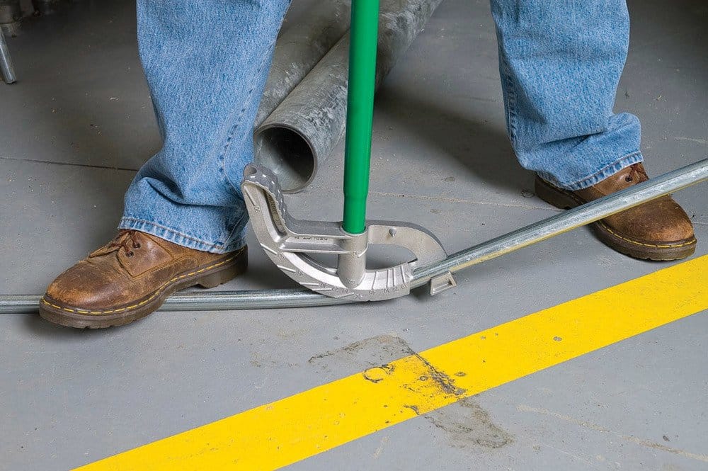 Action shot of a worker wearing work boots using the bender on a concrete floor to create a bend in a silver conduit pipe.