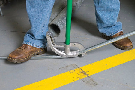 Action shot of a worker wearing work boots using the bender on a concrete floor to bend a silver conduit pipe.