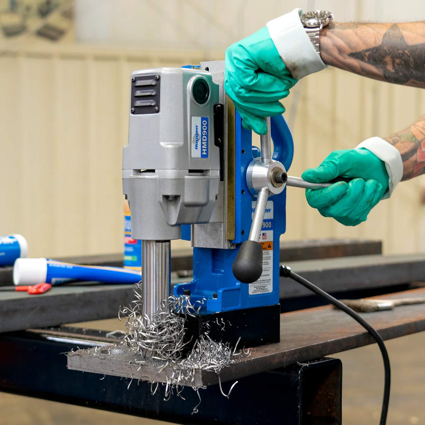 Close-up of gloved hands operating the feed handles of the Hougen HMD900 drill with metal chips accumulating on the base.