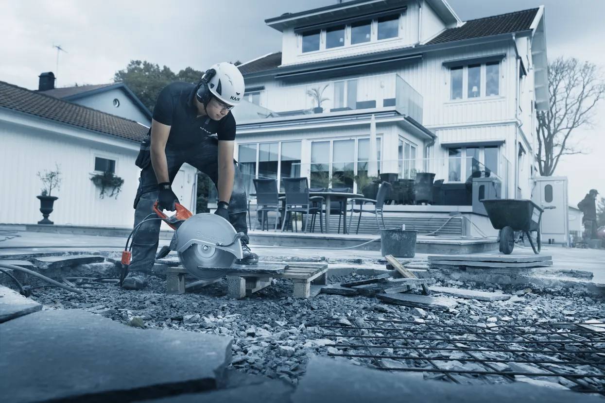  Construction worker using a large circular saw to cut concrete slabs at a residential jobsite with safety gear on.