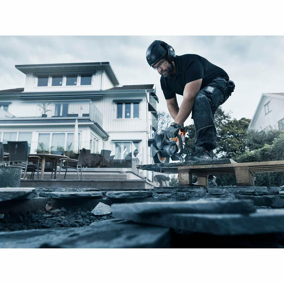 A construction worker wearing ear protection uses the Husqvarna K4000 electric power cutter to cut a dark, flat piece of masonry or paving stone on a wooden pallet outdoors, with a white modern house in the background.