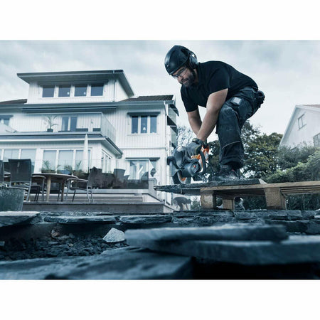 A construction worker wearing ear protection uses the Husqvarna K4000 electric power cutter to cut a dark, flat piece of masonry or paving stone on a wooden pallet outdoors, with a white modern house in the background.