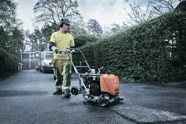 A construction worker uses a Husqvarna LF75 plate compactor on a paved surface near residential homes, wearing full safety gear.