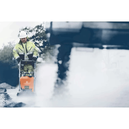 A worker in reflective gear uses a Husqvarna LF75 plate compactor on a misty road surface, with heavy construction equipment visible in the background.