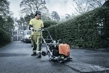 A construction worker operates a Husqvarna LF75 plate compactor on fresh asphalt, with safety gear and a yellow utility vehicle in the background.