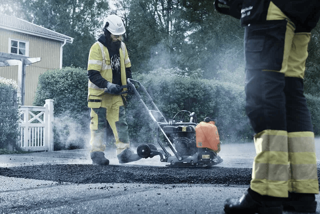 Two construction workers compact hot asphalt using a Husqvarna LF75 plate compactor in a suburban neighborhood, wearing high-visibility safety gear.