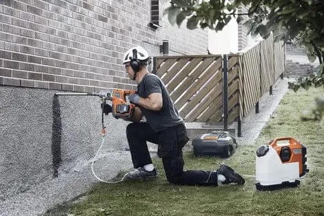 A worker in safety gear kneeling on a lawn uses a handheld core drill connected to the Husqvarna WT15i water tank for dust suppression while drilling into a brick building foundation.