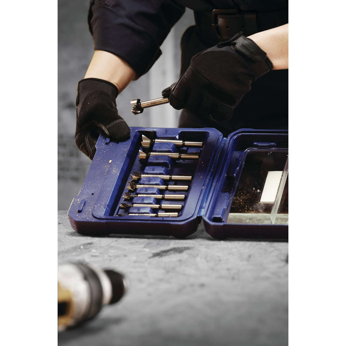 A woodworker wearing protective gloves removes a high carbon steel Forstner bit from the IRWIN Marples 8-piece storage case on a workshop table