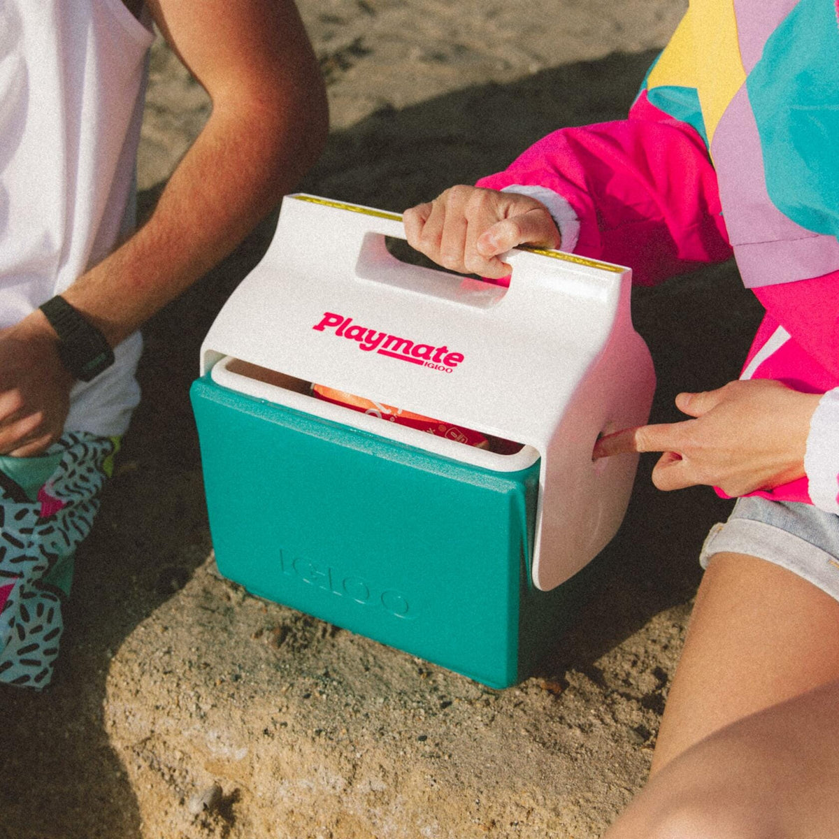 Two people seated outdoors with a teal and white Igloo Playmate Mini cooler between them. One person opens the lid to reveal snacks inside. Cooler displays red “Playmate” text and embossed “Igloo” logo.