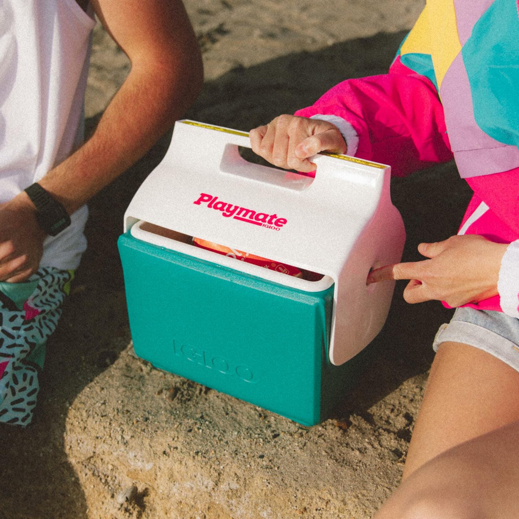 Two people seated outdoors with a teal and white Igloo Playmate Mini cooler between them. One person opens the lid to reveal snacks inside. Cooler displays red “Playmate” text and embossed “Igloo” logo.