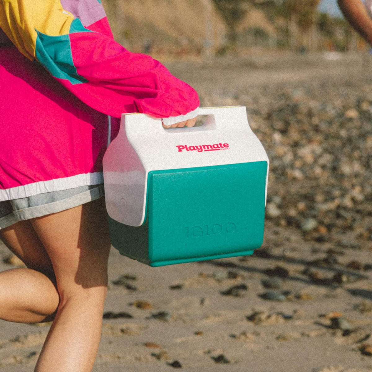 Person walking on a rocky beach carrying a green and white Igloo Playmate Mini cooler. Cooler features red “Playmate” logo and embossed “Igloo” branding. Individual wears a colorful windbreaker and gray shorts.