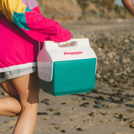 Person walking on a rocky beach carrying a green and white Igloo Playmate Mini cooler. Cooler features red “Playmate” logo and embossed “Igloo” branding. Individual wears a colorful windbreaker and gray shorts.