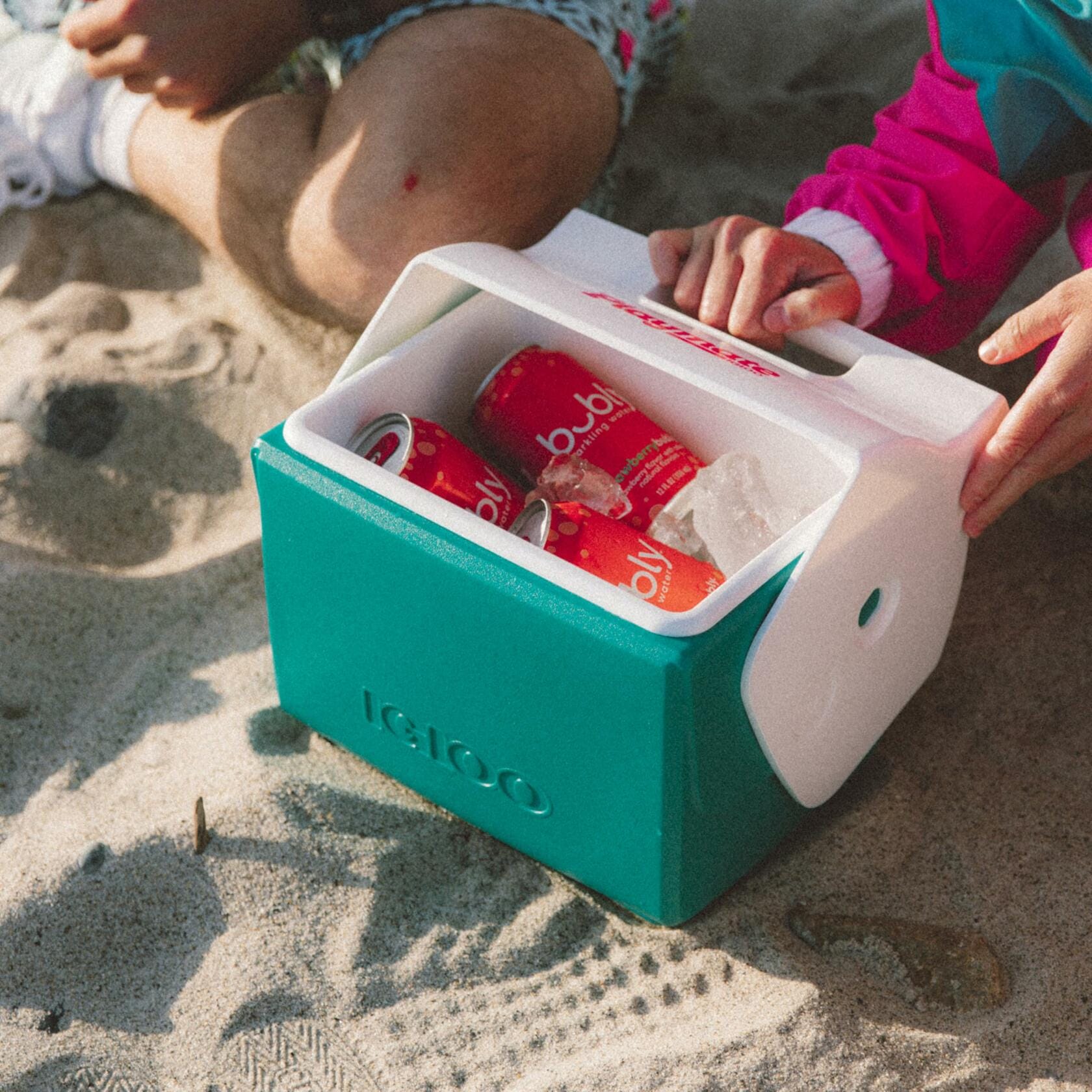 Teal and white Igloo Playmate Mini cooler open on sandy ground with red bubly cans and ice inside. Two people in beachwear reach toward the cooler, suggesting a casual outdoor setting.
