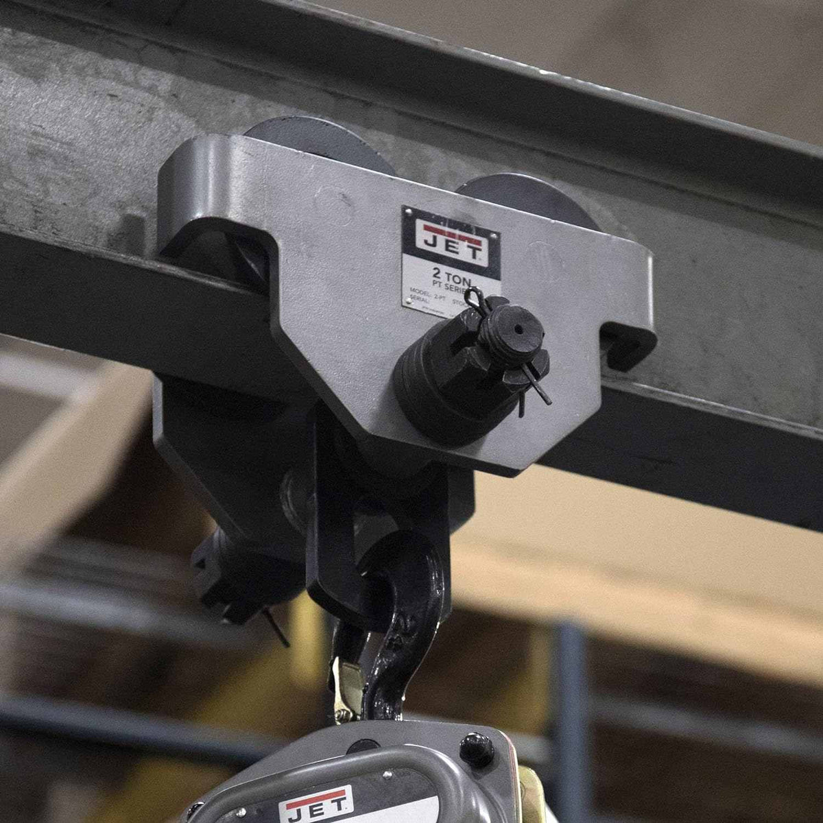 Close-up application shot of a Jet beam trolley securely mounted on an overhead steel I-beam, shown supporting a hoist hook to demonstrate industrial lifting use