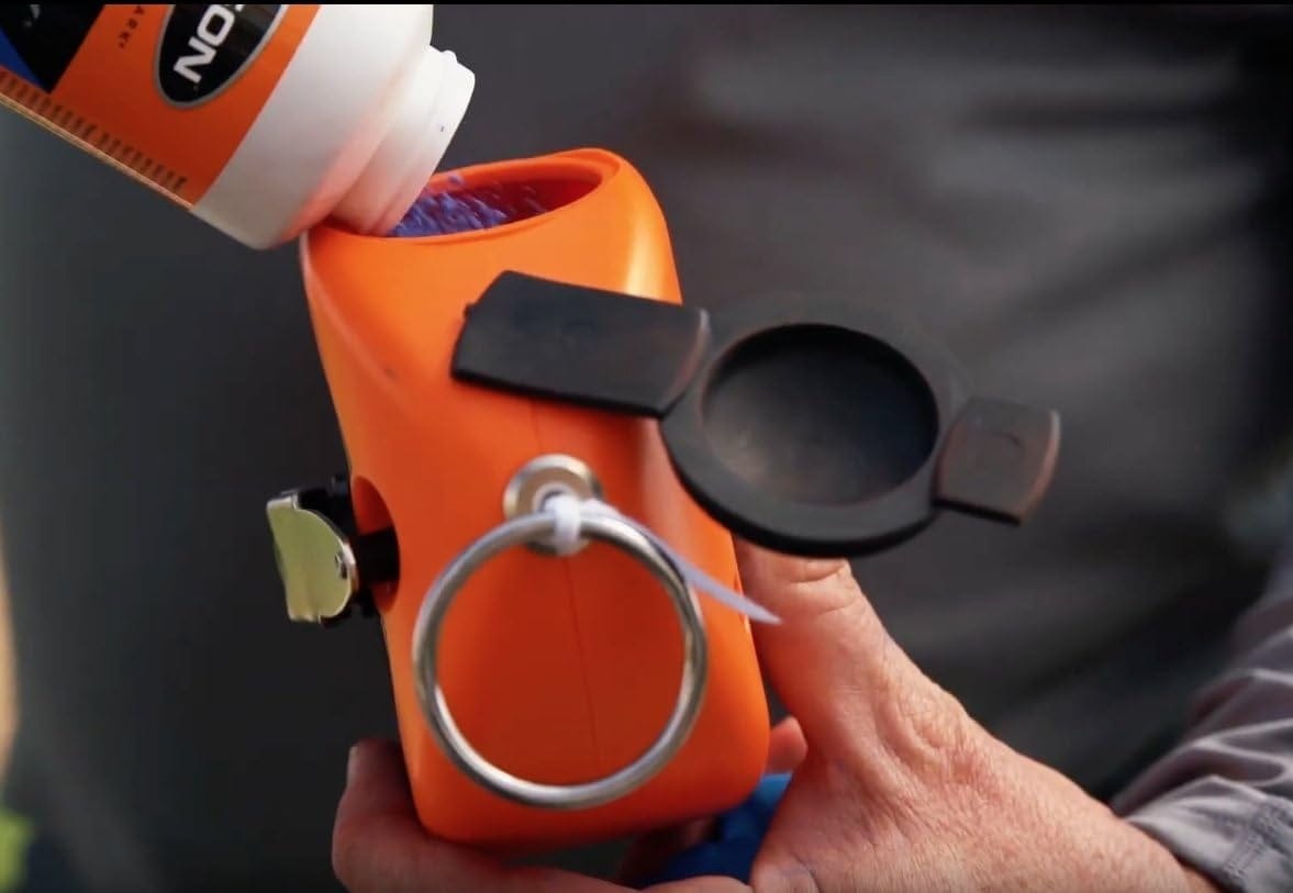 A close-up action shot showing a person pouring blue marking chalk from a bottle into the large BigMouth refill port of an orange Keson Little Giant chalk reel.