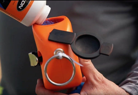 A close-up action shot showing a person pouring blue marking chalk from a bottle into the large BigMouth refill port of an orange Keson Little Giant chalk reel.