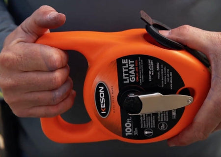 A person's hands holding the orange Keson Little Giant chalk reel, showcasing the shovel-style glovable grip and the flush-mounted steel rewind crank.