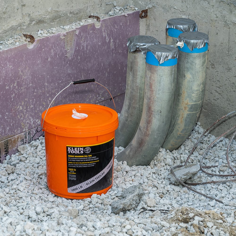 Klein Tools conduit measuring pull tape bucket sitting next to rigid metal elbow conduits on a jobsite ready for commercial cable installation.