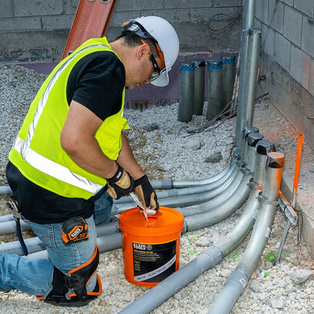 Electrician feeding pre-lubricated 2500-lb pull tape directly from the dispensing bucket into underground PVC conduit stub-ups on a jobsite.