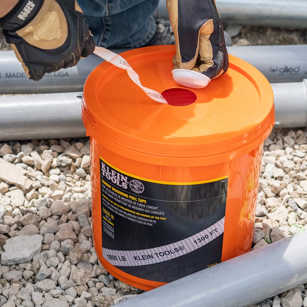 Close-up of a worker pulling white pre-lubricated measuring tape through the bucket lid access aperture on a gravel construction site.