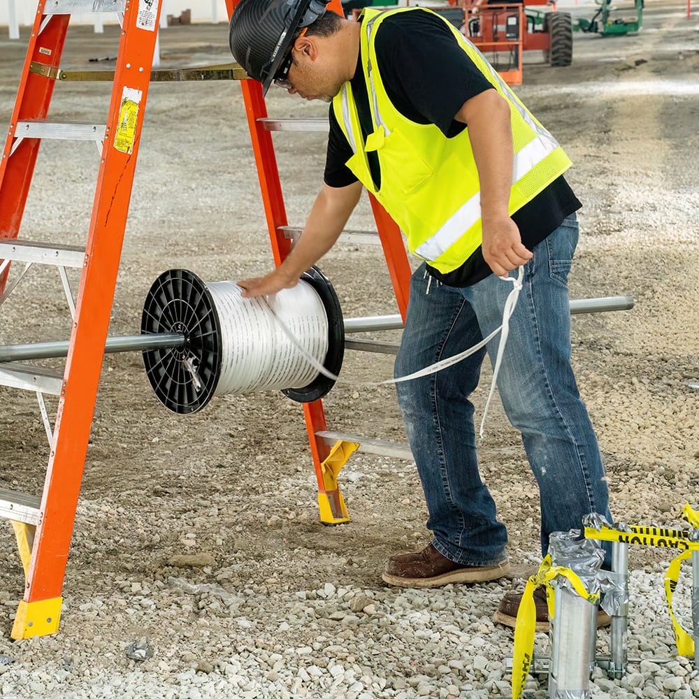 Electrician dispensing pre-lubricated 2500-lb pull tape off a rod-mounted reel setup into underground conduit stub-ups on a jobsite.