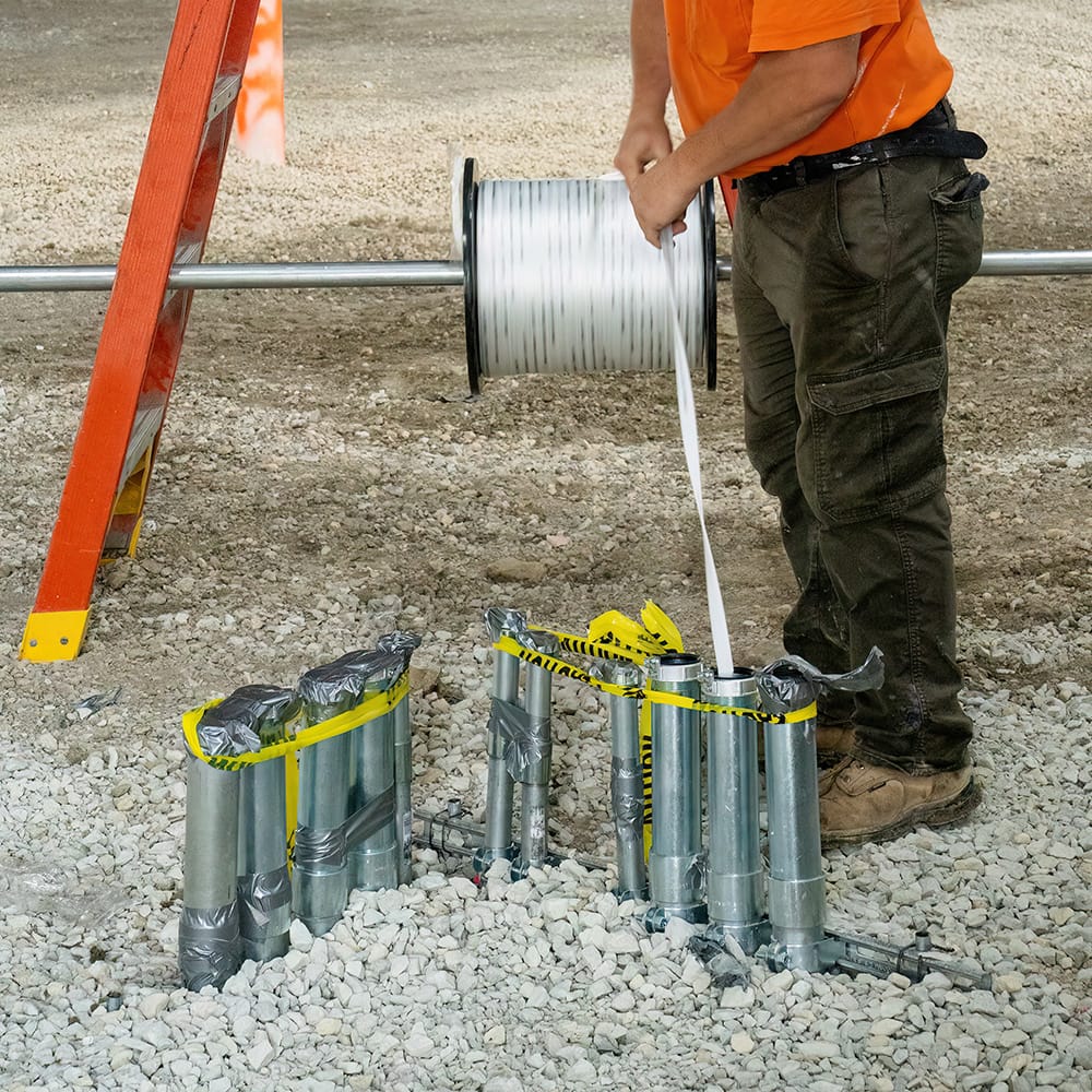 Worker feeding white polyester pull tape from a spindle-mounted reel into PVC underground utility conduits during site construction.