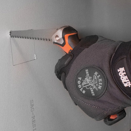 A gloved hand holding the Klein folding jab saw to begin a rectangular cutout in a sheet of drywall.