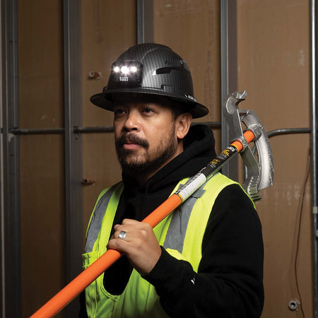 Electrician wearing the illuminated Klein Tools vented hard hat and carrying a conduit bender on a commercial site.