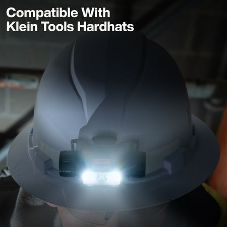 Illuminated Klein Tools 56078 rechargeable LED headlamp securely mounted onto a safety hard hat for hands-free industrial work.