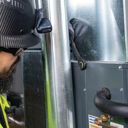 Close-up of a worker in safety glasses and hardhat using the KNECT Pass-Through Ratchet to work on a fastener in a tight space on an HVAC or furnace unit next to a metal pipe.