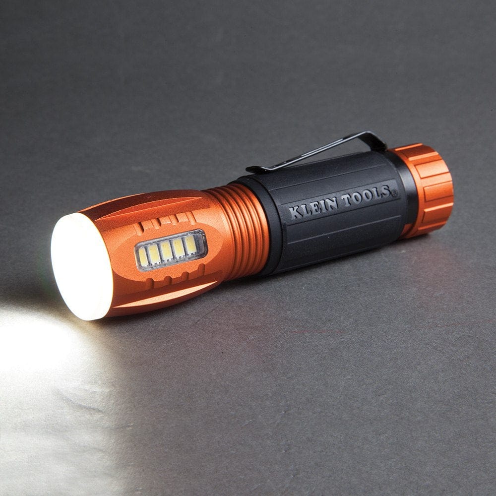 A studio shot of the Klein 56028 flashlight lying on its side, with its main flashlight beam turned on and shining a bright white light.