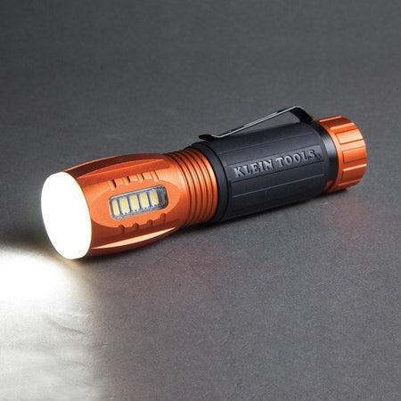 A studio shot of the Klein 56028 flashlight lying on its side, with its main flashlight beam turned on and shining a bright white light.
