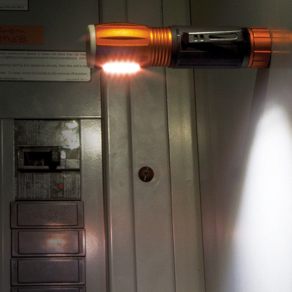 The Klein 56028 flashlight is shown magnetically attached to the front of a metal electrical panel, illuminating the breakers with its side-mounted work light.