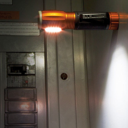The Klein 56028 flashlight is shown magnetically attached to the front of a metal electrical panel, illuminating the breakers with its side-mounted work light.