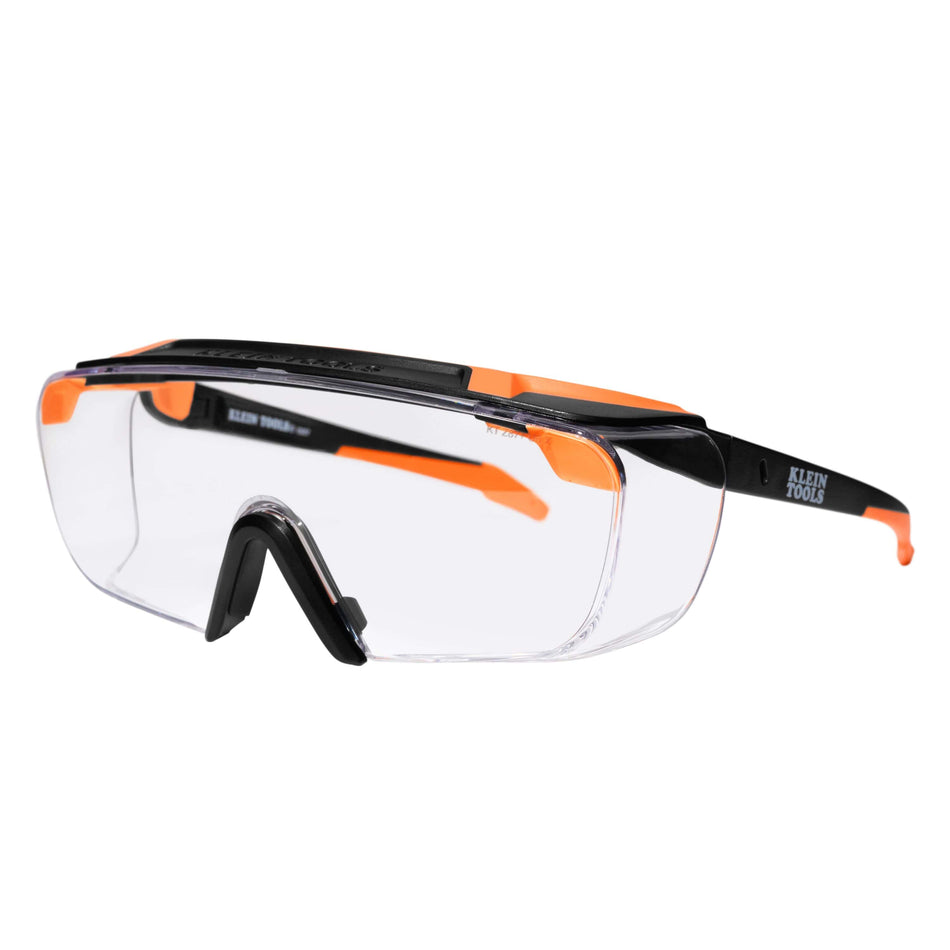 Perspective view of the Klein Tools 03006 over-the-glasses safety glasses featuring a clear high-impact polycarbonate lens, black and orange semi-frame design, and rubberized components for a non-slip fit.