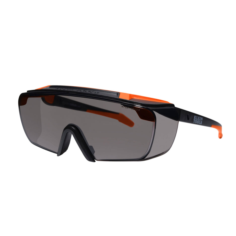 Perspective view of the Klein Tools 03007 over-the-glasses safety glasses featuring gray shaded high-impact polycarbonate lenses, a black and orange nylon semi-frame, and a rubberized nose piece.