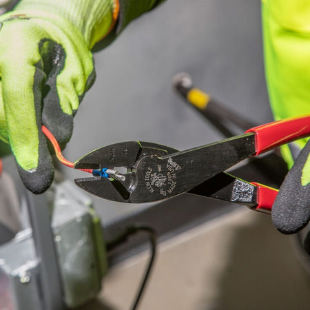 An action shot of a professional wearing high-visibility safety gloves using a Klein 1005 tool to securely crimp a blue insulated terminal onto a red electrical wire near a junction box.