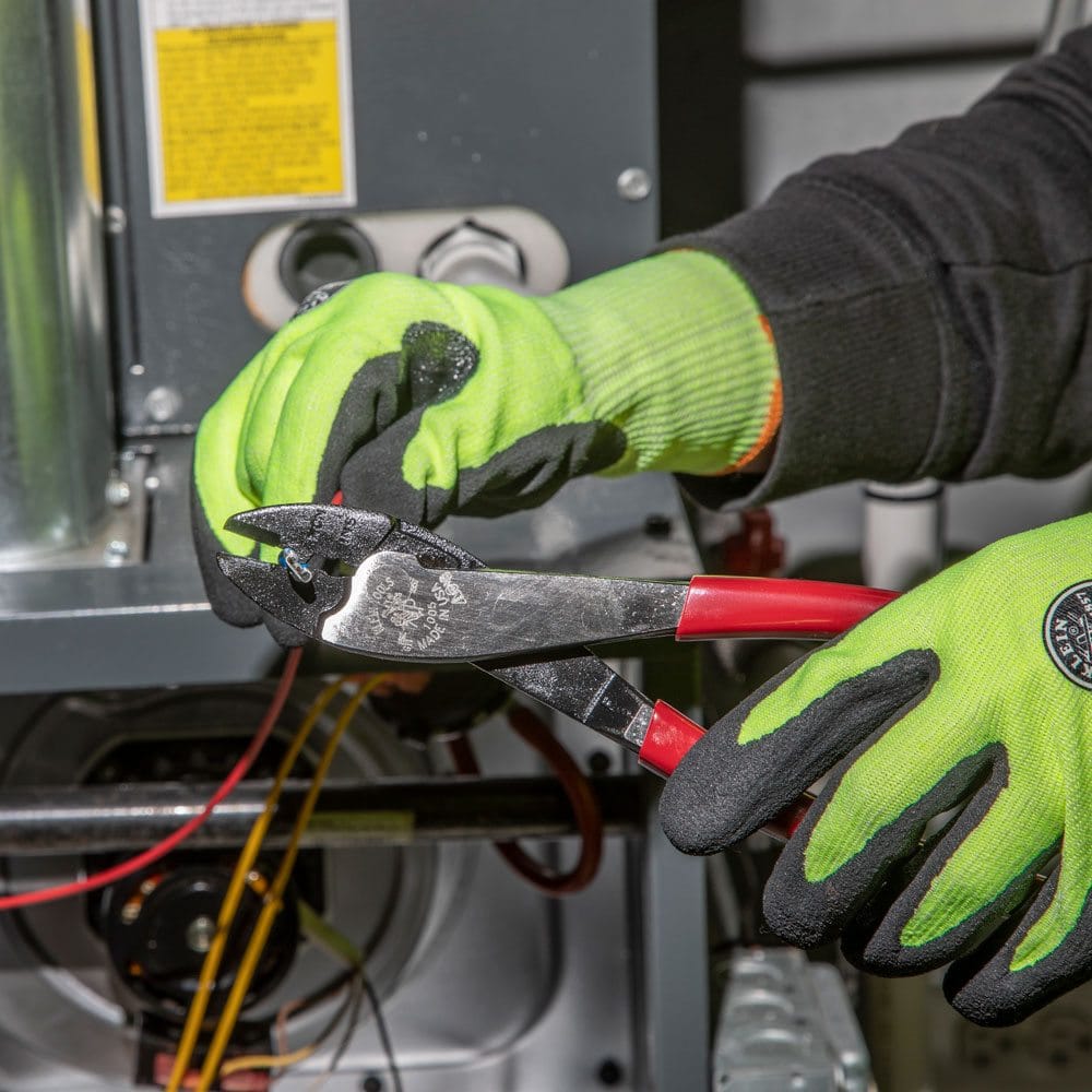 A tradesperson in a work environment using the Klein 1005 9-inch crimper and cutter to perform precise electrical maintenance on a furnace control system, demonstrating its reliability in technical applications.