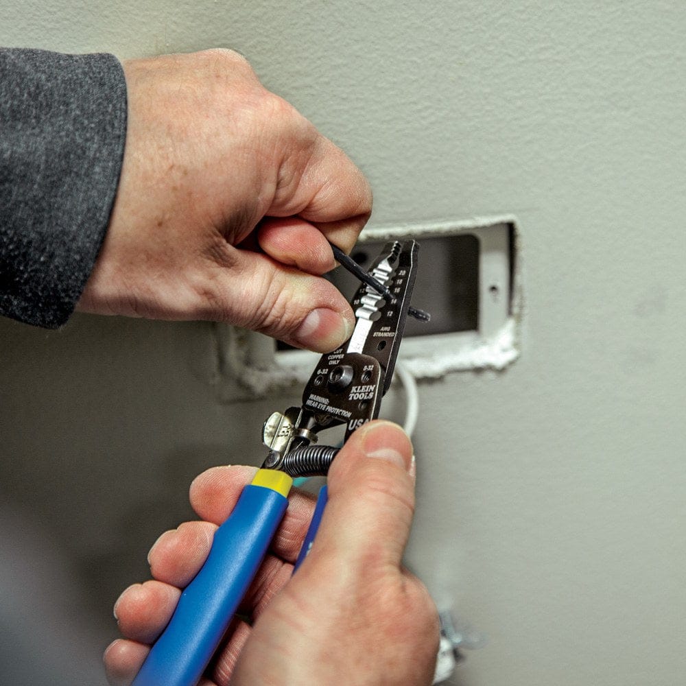 Close-up of hands using the wire stripper to remove insulation from a black wire inside a wall-mounted electrical box.