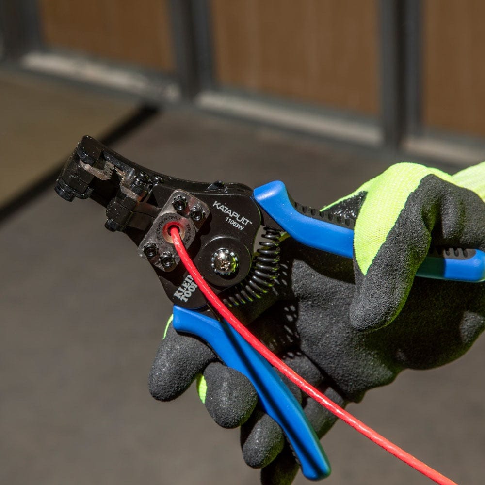 A close-up of a gloved hand using the built-in wire cutter of the Klein Tools Katapult to shear a red wire.