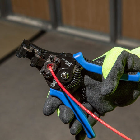 A close-up of a gloved hand using the built-in wire cutter of the Klein Tools Katapult to shear a red wire.