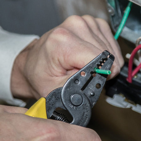 Action shot of a professional using the Klein Tools 11045 wire stripper to accurately strip insulation from a green solid copper wire within an electrical junction box.